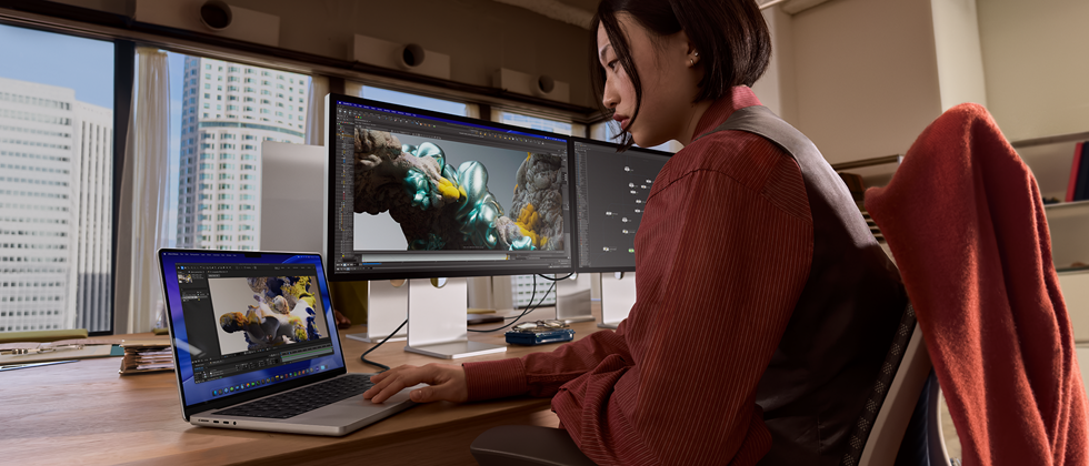 En person sitter på et kontor og jobber på en MacBook Pro som er koblet til Studio Display XDR. Det er høye bygninger utenfor vinduene.