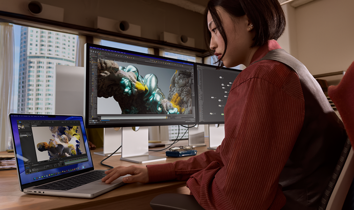 En person sitter på et kontor og jobber på en MacBook Pro som er koblet til Studio Display XDR. Det er høye bygninger utenfor vinduene.