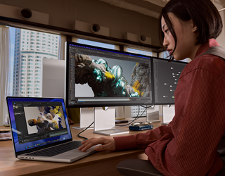 En person sitter på et kontor og jobber på en MacBook Pro som er koblet til Studio Display XDR. Det er høye bygninger utenfor vinduene.