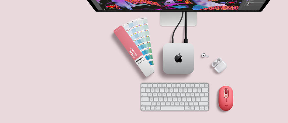 Mac mini vist ovenfra, aluminium i sølv, avrundede hjørner, blank svart Apple-logo i midten, koblet til med kabel, Studio Display er delvis synlig ovenpå. AirPods 4 er åpne, og det vises fargeprøver i Pantone samt et Logitech-tastatur og en Logitech-mus i rødt.